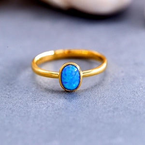 Könnte beinhalten: Ein goldfarbener Ring mit einem zentralen, oval geformten blauen Opal-Cabochon. Das glatte, polierte Metallband hält den Stein in einer einfachen Zargenfassung und erzeugt ein klares und schlichtes Design, das die leuchtend blaue Farbe betont.