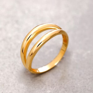 May include: A polished gold ring featuring a double band design. The ring consists of two smooth, rounded, parallel bands that are connected at the sides, creating an open space between the two circular segments with a reflective metallic finish.