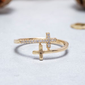 May include: A gold ring featuring two crosses, one with small diamonds, the other plain. The ring is on a white surface.