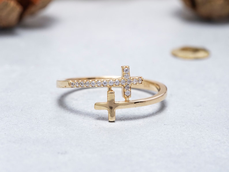 May include: A gold ring featuring two crosses, one with small diamonds, the other plain. The ring is on a white surface.