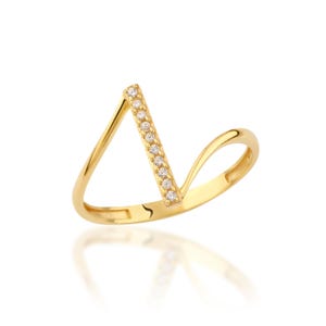 May include: A gold-colored ring with a V-shaped design. The vertical bar of the V is adorned with small, clear gemstones. The ring has a smooth, polished finish and reflects light.