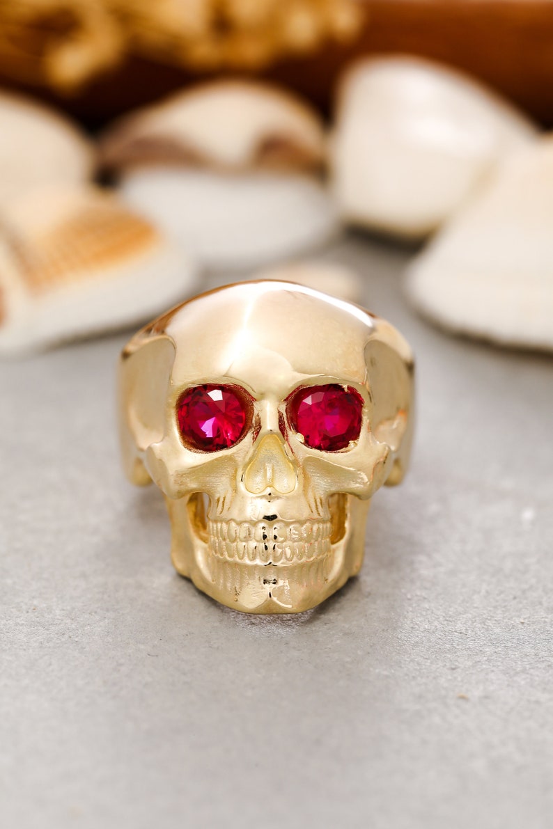 May include: A gold skull ring with red gemstone eyes. The ring is detailed with a skull face and teeth.