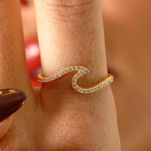 Puede incluir: Un anillo de oro en forma de ola con piedras de circonita cúbica transparentes. El anillo se lleva en un dedo.