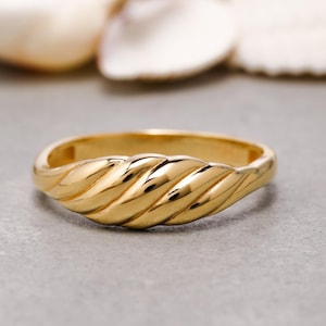 Puede incluir: Un anillo de oro pulido que presenta una banda elevada prominente con una serie de ranuras diagonales, retorcidas o acanaladas en la superficie superior. El metal tiene un acabado liso y reflectante que capta la luz.