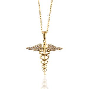 Peut inclure: Un collier doré avec un pendentif Caducée, symbole de la médecine. Le pendentif est orné de deux ailes incrustées de petites pierres précieuses transparentes. La chaîne du collier est également dorée, sur fond blanc.