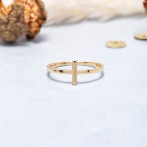 May include: A gold-toned ring featuring a simple cross design. The ring is thin and delicate, with a smooth finish.
