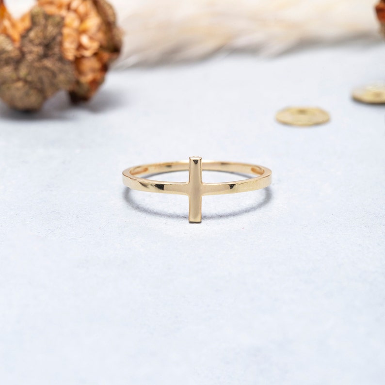 May include: A gold-toned ring featuring a simple cross design. The ring is thin and delicate, with a smooth finish.