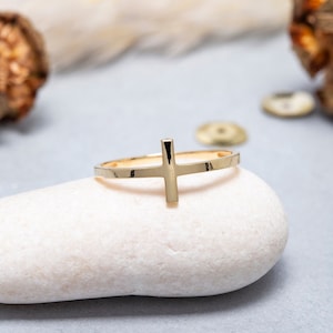 May include: A gold-toned ring with a simple cross design. The ring is resting on a white stone.