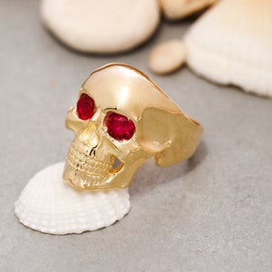 May include: A gold skull ring with red gemstone eyes. The ring is sitting on a white seashell.