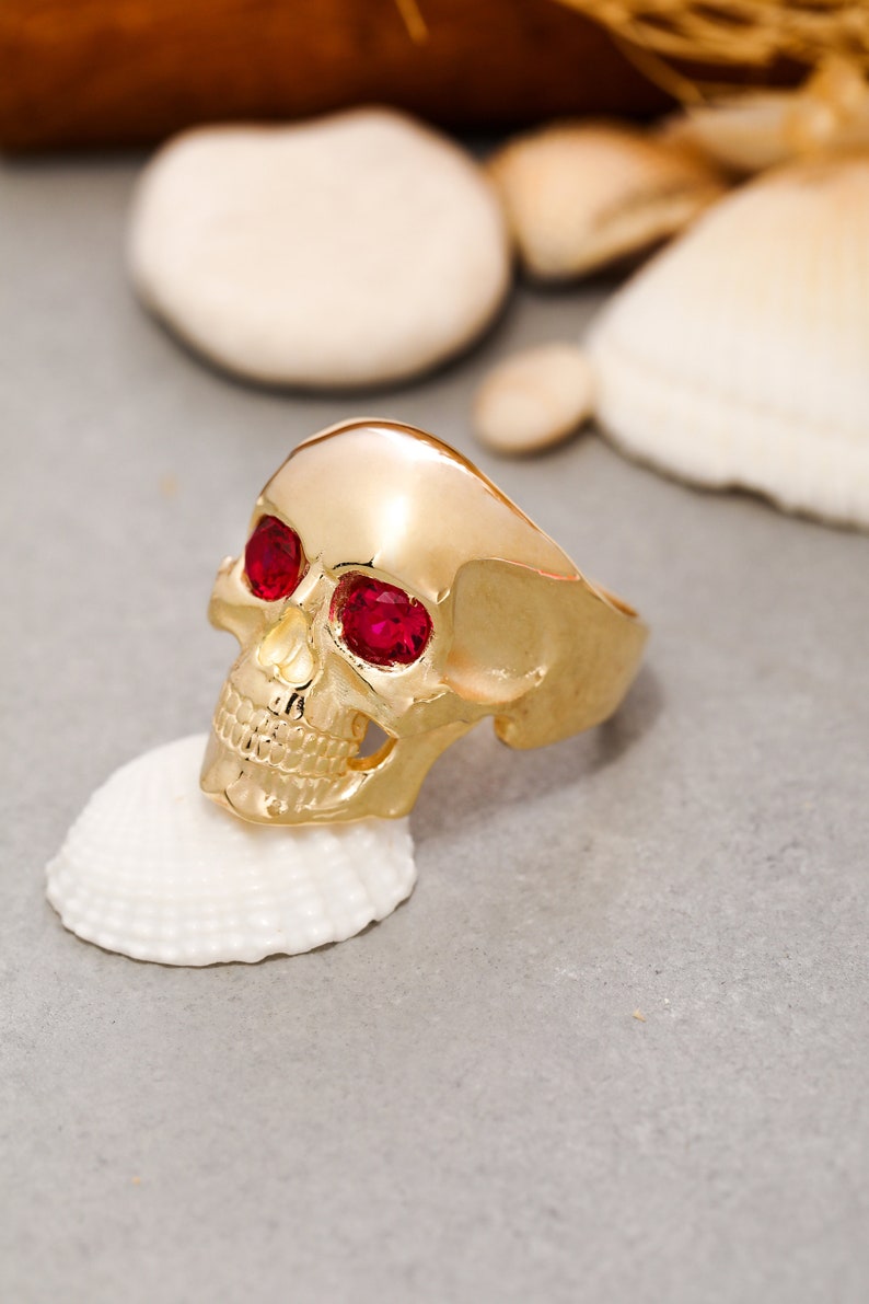 May include: A gold skull ring with red gemstone eyes. The ring is sitting on a white seashell.