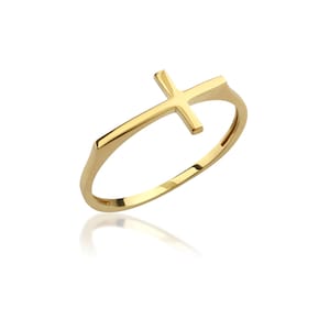 Puede incluir: Un anillo de oro pulido que presenta un diseño de cruz horizontal integrado en la banda. El anillo tiene un acabado metálico liso y reflectante con una estructura simple y limpia. La cruz está colocada en la parte superior de la banda circular, creando una pieza de joyería continua y unificada con un tono de oro amarillo brillante.