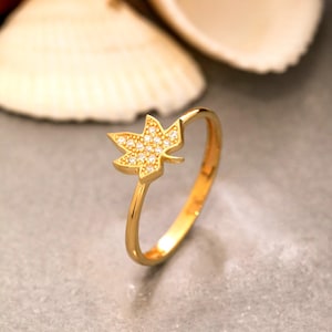 Può includere: Un anello in oro con un elemento centrale a forma di foglia d'acero decorato con piccole pietre chiare e scintillanti. Il gambo è sottile e lucido, caratterizzato da una finitura metallica liscia e riflettente che cattura la luce su tutta la struttura.