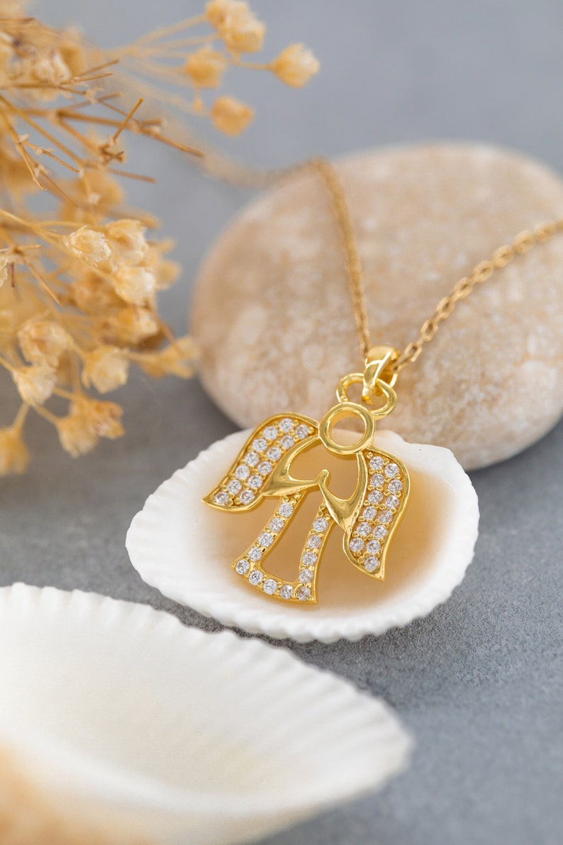 K&ouml;nnte beinhalten: Eine goldene Engels-Anh&auml;nger-Kette mit klaren Zirkonia-Steinen. Die Kette wird auf einer wei&szlig;en Muschel pr&auml;sentiert.