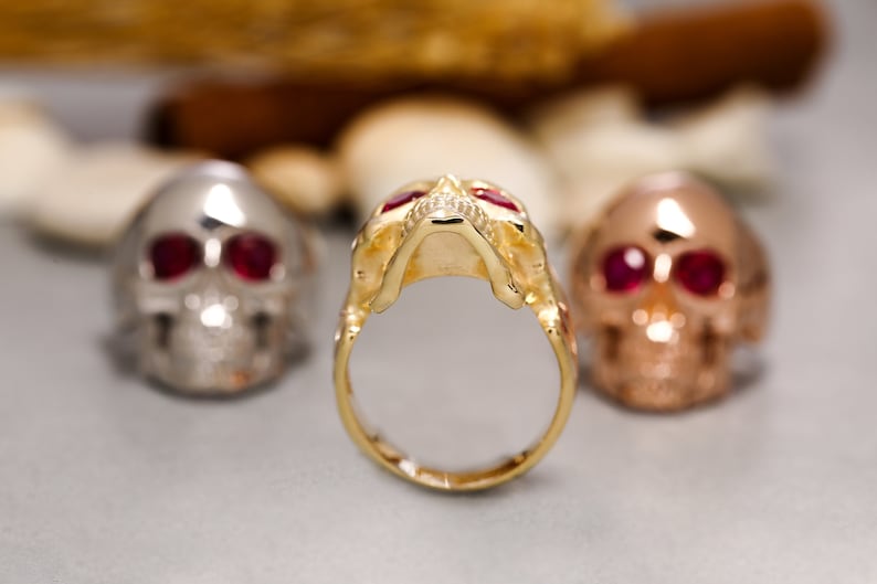 May include: Three gold skull rings with red gemstone eyes. The ring in the centre is in focus and the other two are out of focus.