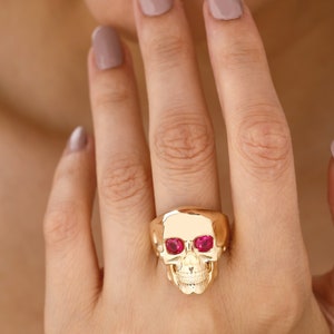 May include: A gold skull ring with ruby red eyes. The ring is worn on a finger with light pink nail polish.
