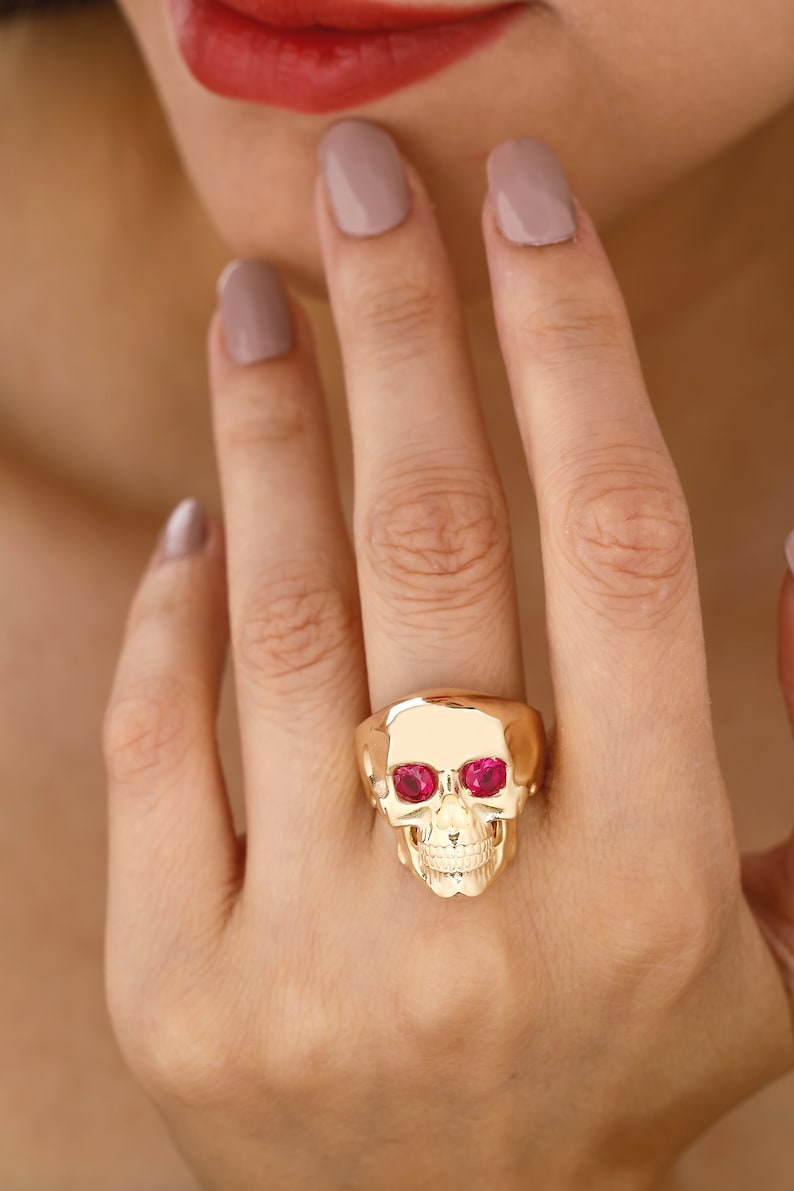 May include: A gold skull ring with ruby red eyes. The ring is worn on a finger with light pink nail polish.