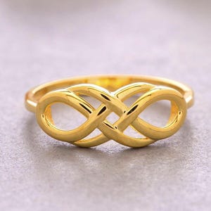 May include: A polished gold ring featuring a central infinity symbol design. The band is smooth and circular, connecting to the intertwined loops that form the horizontal figure-eight shape, creating a continuous and reflective metallic surface.