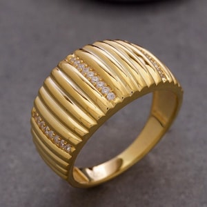 Könnte beinhalten: Ein breiter Goldring mit einer Reihe vertikaler, erhabener Rippen auf der Oberfläche. Zwei parallele Reihen kleiner, klarer, rund geschliffener Steine sind in die gerillte Textur eingebettet und erzeugen ein reflektierendes, metallisches Aussehen.