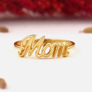 Puede incluir: Un anillo de tono dorado que presenta la palabra Mom en una fuente de escritura cursiva. La superficie metálica es pulida y reflectante, mostrando una banda lisa y continua que se conecta con las letras. El diseño está centrado y es legible.