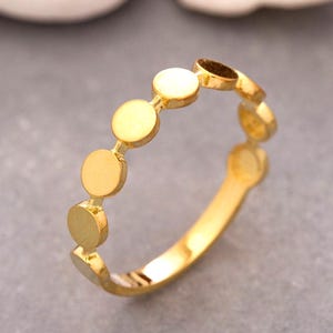 Könnte beinhalten: Ein goldfarbener Ring mit einer Reihe kleiner, flacher, kreisförmiger Scheiben, die entlang der Oberseite des Bandes angeordnet sind. Das Metall hat eine polierte, reflektierende Oberfläche und die kreisförmigen Elemente bilden ein durchgehendes Band.