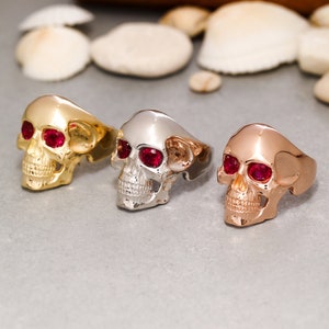 May include: Three skull rings with red gemstone eyes. The rings are made of gold, silver, and rose gold.