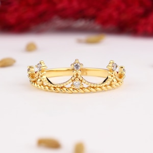 Può includere: Un anello a corona color oro con un design a corda intrecciata. L'anello presenta piccole gemme trasparenti incastonate sulle punte e al centro della corona. L'anello è su una superficie bianca con uno sfondo rosso sfocato.