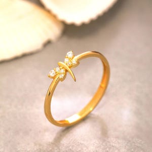 Può includere: Un anello in oro caratterizzato da un delicato motivo a libellula sulla fascia. La libellula è impreziosita da piccole pietre chiare e scintillanti incastonate nelle ali e nel corpo, creando un aspetto raffinato sulla sottile fascia in oro.
