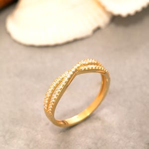 Pode incluir: Um anel de ouro com um design cruzado de duas bandas delicadas que se encontram no centro. A superfície das bandas é incrustada com pequenas pedras claras e brilhantes que captam a luz, criando um aspeto refinado e polido.