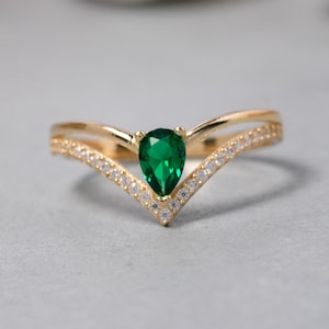May include: A gold ring featuring a pear-shaped emerald gemstone and a V-shaped band adorned with small clear stones.