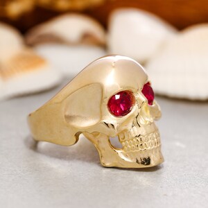 May include: A gold skull ring with red gemstone eyes. The ring is detailed with a skull face and teeth.
