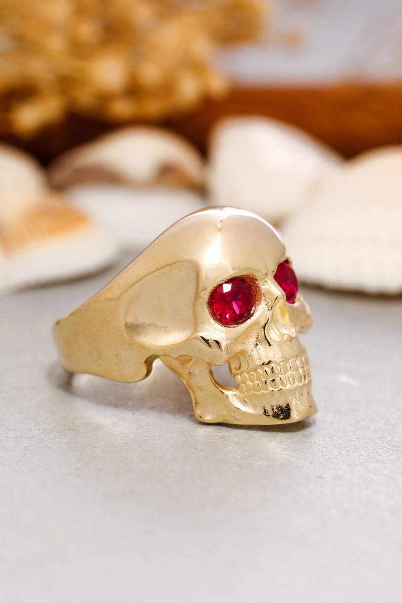 May include: A gold skull ring with red gemstone eyes. The ring is detailed with a skull face and teeth.