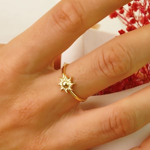 May include: A gold-toned ring with a sun design. The ring is on a person's finger.
