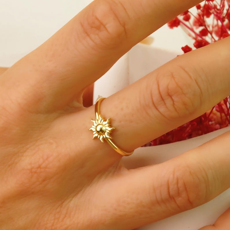 May include: A gold-toned ring with a sun design. The ring is on a person's finger.