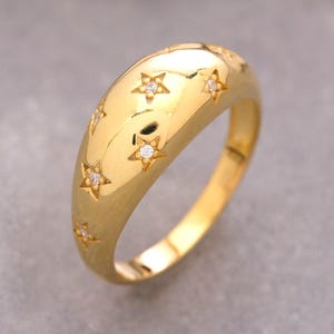 Könnte beinhalten: Ein polierter Goldring in Kuppelform mit einer Reihe eingravierter Sternmotive auf der Oberfläche. Jeder Stern ist mit einem kleinen, klaren, funkelnden Edelstein in der Mitte versehen, was ein himmlisches Muster auf dem glatten Band erzeugt.