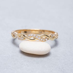 Könnte beinhalten: Ein goldener Ring mit einem gedrehten, geflochtenen Banddesign. Eine Hälfte des Bandes besteht aus poliertem Gold, während die andere Hälfte mit kleinen, klaren, rund geschliffenen Steinen besetzt ist, die das Licht einfangen und funkeln.