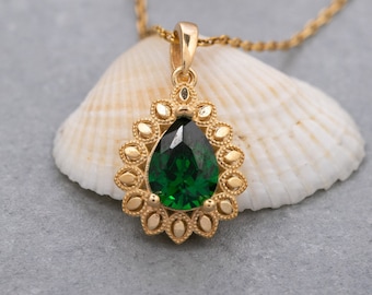 May include: A gold necklace with a pear-shaped green gemstone pendant surrounded by a decorative gold filigree design. The necklace is resting on a white seashell.