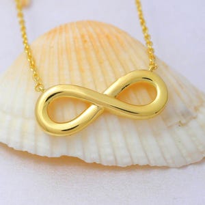 May include: A gold-toned infinity symbol pendant suspended from a delicate matching chain. The smooth, polished metal infinity charm features a continuous loop design and is positioned against a textured, off-white seashell background.