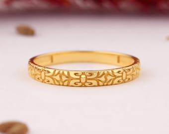 May include: A gold-colored ring with an intricate, repeating floral pattern. The ring is set against a soft, neutral background, highlighting its detailed design. The ring's surface reflects light, showcasing its polished finish.