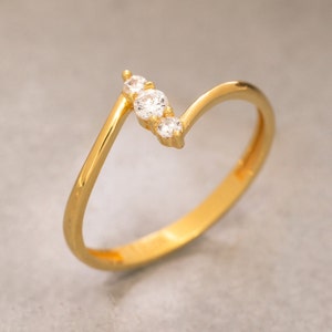 May include: A gold-colored ring with a wave design. The ring features three small, clear gemstones set in a row. The band is thin and smooth, with a polished finish. The ring is set against a neutral background.