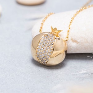May include: Gold-colored pomegranate pendant necklace. The pendant features a rounded shape with a band of small, sparkling stones. The necklace hangs on a delicate gold chain, creating an elegant and eye-catching piece of jewelry.