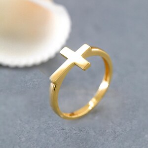 Peut inclure: Une bague en or poli présentant un motif de croix proéminent intégré à l'anneau. La bague possède une surface lisse et réfléchissante ainsi qu'une structure simple et épurée, mettant en valeur une finition métallique classique et lumineuse.