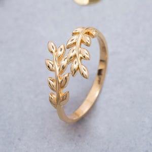May include: A gold ring featuring an open bypass design with two overlapping branches. Each branch is adorned with small, polished gold leaves that extend along the band, creating a continuous botanical motif in a bright, reflective metallic finish.