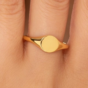 May include: A polished gold signet ring featuring a smooth, circular face and a tapered band. The ring has a reflective, metallic finish and a simple, clean design with no engravings or stones, showcasing a solid gold construction throughout.