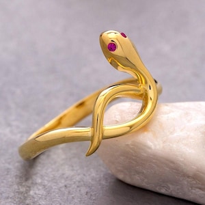 May include: A gold-toned ring shaped like a coiled snake with a smooth, polished finish. The snake head features two small, vibrant pink circular stones set as eyes. The band curves to form the body of the serpent, creating a continuous, sleek loop.