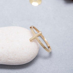 May include: A gold-toned ring with a simple cross design. The ring is resting on a white stone.