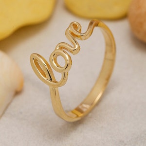 May include: A polished gold-tone ring featuring the word love written in a continuous cursive script as the focal point. The band is smooth and circular, connecting to the base of the letters. The metal has a reflective, shiny finish.