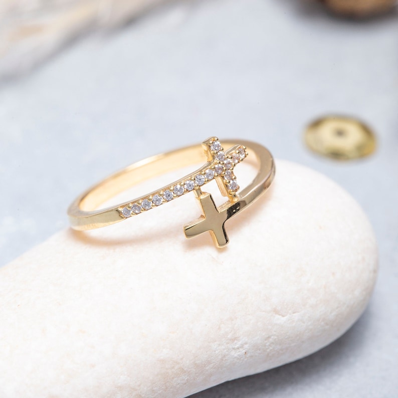 May include: A gold ring with two crosses, one with a row of small clear stones. The ring is on a white stone.