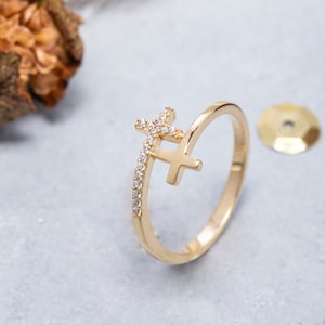 May include: A gold ring with a delicate cross design featuring small clear stones. The cross is set on a thin band with a row of stones along the side.