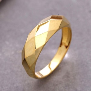 May include: A gold ring featuring a faceted, geometric surface with multiple angled planes. The band has a polished, reflective finish that catches the light, creating alternating bright and shadowed sections across its textured, multi-sided exterior.