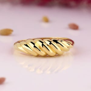 Könnte beinhalten: Ein Ring aus poliertem Gold mit einem durchgehenden, abgerundeten, gedrehten Seildesign, das sich um das gesamte Band zieht. Die Oberfläche des Metalls ist stark reflektierend und erzeugt deutliche Glanzlichter und Schatten entlang der Kanten.